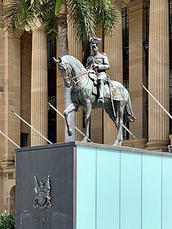 Equestrian statue in dark grey metal of George V in military dress uniform on a plinth of red granite outside a Classical building of red sandstone