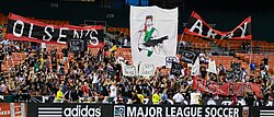 Fans wearing black cheer with several large graphics in a stadium's bleachers.