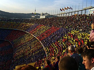 A crowd of spectators in an open-air stadium, a mosiac in red, blue and gold is visible