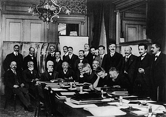 Photograph of the first conference, 2 November, 1911, at the Hotel MetropoleSeated (L–R): W. Nernst, M. Brillouin, E. Solvay, H. Lorentz, E. Warburg, J. Perrin, W. Wien, M. Skłodowska-Curie, and H. Poincaré.Standing (L–R): R. Goldschmidt, M. Planck, H. Rubens, A. Sommerfeld, F. Lindemann, M. de Broglie, M. Knudsen, F. Hasenöhrl, G. Hostelet, E. Herzen, J. H. Jeans, E. Rutherford, H. Kamerlingh Onnes, A. Einstein and P. Langevin.