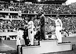 Jesse Owens on the podium after winning the long jump at the 1936 Summer Olympics, where he won four gold medals; in his 1980 obituary, The New York Times called him "perhaps the greatest and most famous athlete in track and field history".[214]