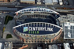 Aerial view of an empty stadium with prominent arches. The surrounding area has railroad tracks, a plaza, and a parking garage.