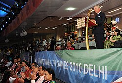 Charles in a suit speaking at a podium during the 2010 Commonwealth Games. Attendees sit around him.