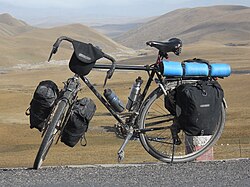 Touring bicycle equipped with front and rear racks, fenders (called mud-guards), water bottles in cages, four panniers and a handlebar bag
