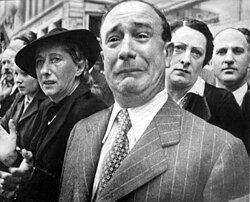 A man in front of a well dressed crowd crying while appearing to be suppressing his emotions by pressing his lips and contorting his forehead