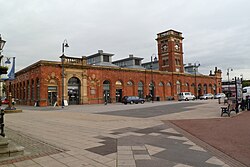 A red-brick, mostly single-storey building as photographed from across a pedestrianised square. There are frequent floor-to-ceiling windows (thus spanning the whole height of the building), with a single cuboidal spire at the centre of one façade extending upwards.