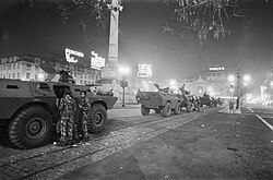 Black-and-white nighttime photograph of several armoured military vehicles lined up in Rossio Square, with soldiers standing beside them near the Column of Pedro IV and bright neon signs glowing in the background.