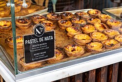 Photograph of a bakery display case filled with pastéis de nata, with a sign in front labelled "Pastel de Nata" and describing them as a low-sugar secret recipe in Portuguese and English.