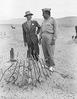 A man smiling in a suit in suit and one in a uniform chat around a pile of twisted metal.