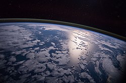 A view of the Southern Pacific under moonlight with different layers of Earth's atmosphere visible: the troposphere with its clouds casting shadows, a band of stratospheric blue sky at the horizon, and a line of green airglow of the lower thermosphere around an altitude of 100 km, at the edge of space