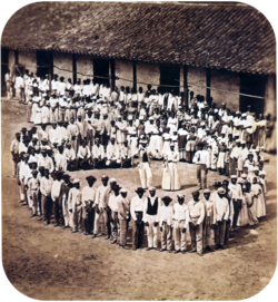 Photograph showing a group of people dressed in white, who have gathered in front of a tile-roofed farm building and observe another large group which has formed a large circle surrounding five men straddling large drums, a woman and two other men