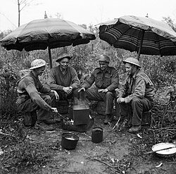 Gunners of the 78th Field Regiment, Royal Artillery make use of two sunshades from a cafe to keep the rain off while making a brew, Anzio, Italy, 27 February 1944.