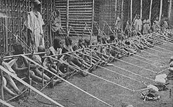 A row of more than a dozen children holding wooden looms stretches into the distance.