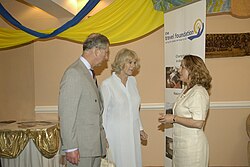 Charles wearing a suit converses with a woman in a dress. Queen Camilla stands next to Charles in the background standing opposite a "Travel Foundation" vertical banner.