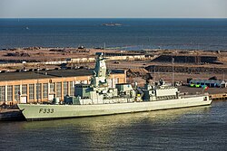 Photograph of NRP Bartolomeu Dias (F333) of the Portuguese Navy docked by the quays of Hernesaari, Helsinki, Finland, with the Portuguese flag flying at the stern and calm blue water stretching out to the open sea beyond.