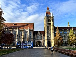 A grand gothic church-like building as viewed from a short distance away across a pedestrianised street. The building has a grand stained-glass window at the front surrounded by two cuboid towers, and a large cuboid tower at the back-right with a red brick roof.