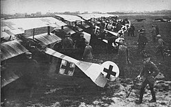 A black and white image of triplanes lined up on grass a black Iron Cross on the back of each of them with many people behind them