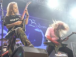 A color photograph of two members of the group Children of Bodom standing on a stage with guitars, drums are visible in the background. Both electric guitarists have "flying V" style guitars and they have long hair.