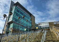 A glass and steel building on the left of a photograph, as viewed from the bottom of a large set of outdoor stairs leading up to it. The view of the building is partially obscured by lamp posts and the stairs are split up by steel railings.
