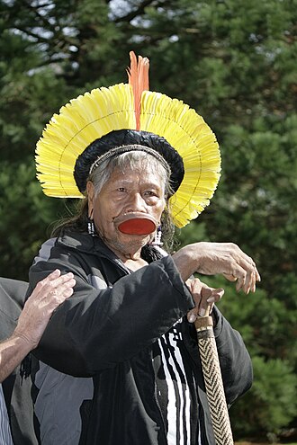 An elderly man is looking forward. He wears traditional garments on his head and lower lip