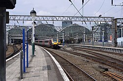 An aerial photo of a modern railway terminus station viewed from the top end a short distance away. The multiple parallel roofs of the trainshed stretch into the background against a grey sky.