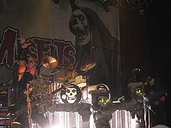 The rock band The Misfits performing onstage. The band's name in large lettering is printed on a fabric panel behind the performers along with a skull image. From left to right are the electric bassist, drummer, and electric guitarist.