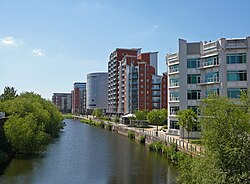 The canalised River Aire flows from the Dark Arches under Leeds main railway station towards the bottom of the picture. To the left of the river is the lock which links the river with the Leeds and Liverpool Canal. To the right is a riverside walk beneath modern buildings, and in the distance, beyond the railway viaduct and station, are more high-rise modern buildings located on the west side of the city centre.
