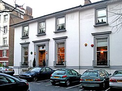 A number of cars are parked in the asphalt car-park of a two-storey white building. The building appears once to have been detached, but a two-storey extension is apparent, on the right side of the building. The ground floor contains three large sash windows, each with a small metal railing around the sill. The first floor contains four shorter sash windows.  The decorative stonework around the windows and main entrance has been painted grey. Two chimney stacks are visible on the roof, at opposite ends. The staircase entrance to the building is bordered by metal railings. To the left of the image, a much larger brick building can be seen in the distance.