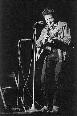 Dylan with his guitar onstage, laughing and looking downwards.
