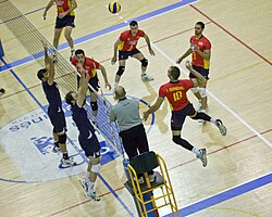 A Spanish player, #18 in a red outfit is shown about to spike towards the Portuguese field, whose players are performing a block