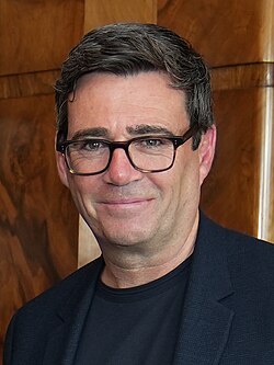 A fairly young man slightly smiling at the camera against green shrubbery as a background. He is wearing square glasses, a blue tie, and a grey blazer on a white shirt.