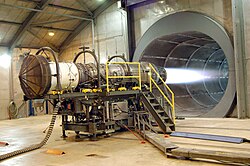 A fighter jet engine undergoing testing. The tunnel behind the engine allows noise and exhaust to escape.