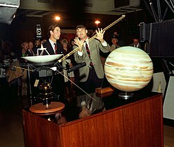 Charles and another man dressed in a suit discuss space exploration beside models of a spacecraft and Jupiter as a crowd watches behind them.