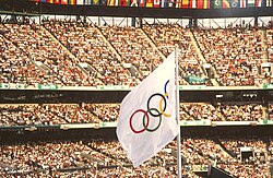 A white flag with five differently colored interlocking rings in the center is seen atop a flagpole, against the backdrop of a stadium filled with spectators.