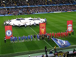 The Chelsea (blue) and Liverpool (red) teams line up side-by-side in front of banners bearing their club crests and the Champions League final logo. In the background, a flag bearing the Champions League logo is waved over the centre circle, while the main stand is further back, full of spectators. In the foreground, Chelsea fans wave blue flags bearing their club crest.