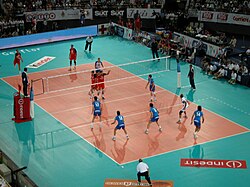 An international match between Italy and Russia in 2005; the Russian player on the left has just served with three men from his team next to the net moving to their assigned block positions from the starting ones. Two others, in the back-row positions, are preparing for defense. Italy, on the right, has three men in a line, each preparing to pass if the ball reaches him. The setter is waiting for his pass while the middle hitter (#10) will jump for a quick hit if the pass is good enough. Alessandro Fei (#14) has no passing duties and is preparing for a back-row hit on the right side of the field.