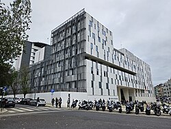 Photograph of the headquarters of the Judicial Police (PJ). A large modern white-and-grey office building with irregular window patterns stands on a corner under an overcast sky, with parked motorcycles, a few pedestrians, and a crosswalk in the foreground.