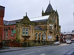 The façade of Ashton town library, constructed from stone and built in Gothic revival style. The photo is a colour modern photo taken from across the road.