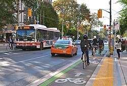 A roadway with bike lanes. Orion VII operated by the Toronto Transit Commission is visible in the background.