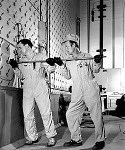 Two workmen in overalls put a rod into a hole on the reactor face