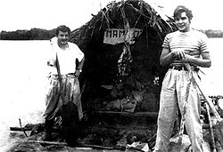 black and white photograph of two men on a raft, fitted with a large hut. The far bank of the river is visible in the far distance