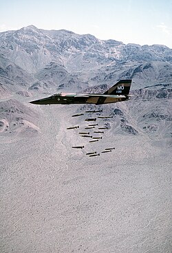 An F-111A drops 24 Mark 82 low-drag bombs in-flight over a bombing range.