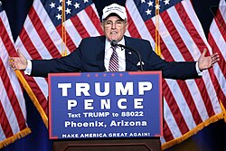 Giuliani with his arms outstretched, standing a podium with a banner for the Trump/Pence ticket