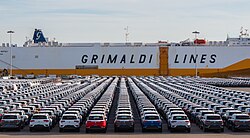 Rows of newly manufactured cars parked at a port in front of a Grimaldi Lines vehicle carrier.