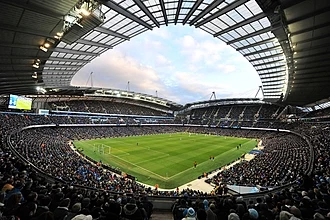 A football stadium as viewed from one corner. The photograph is a fish-eye panorama taken during a game against a blue sky. The stadium is full of fans on all four sides of the pitch, with large stands stretching up below an arched roof.