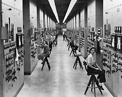 A long corridor with many consoles with dials and switches, attended by women seated on high stools
