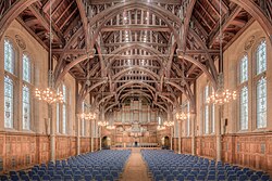The interior of a grand hall as photographed from the back-centre. The hall is full of chairs and has a grand and intricate beamed roof with chandeliers hanging from the beams and gothic-shaped windows either side.
