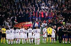 A line of men in white shirts watching men in blue-and-red shirts standing on a rostrum in the background.