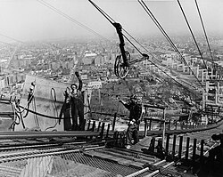 Two men are standing high in the air on a walkway, and a wheel is above them, suspended by wires.