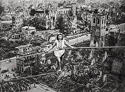 A black and white picture of a woman, wearing a blouse and shorts, holding a long stick with her two hands to balance herself while walking on a tightrope, above ruins of buildings and a church. People form a crowd on the ground.